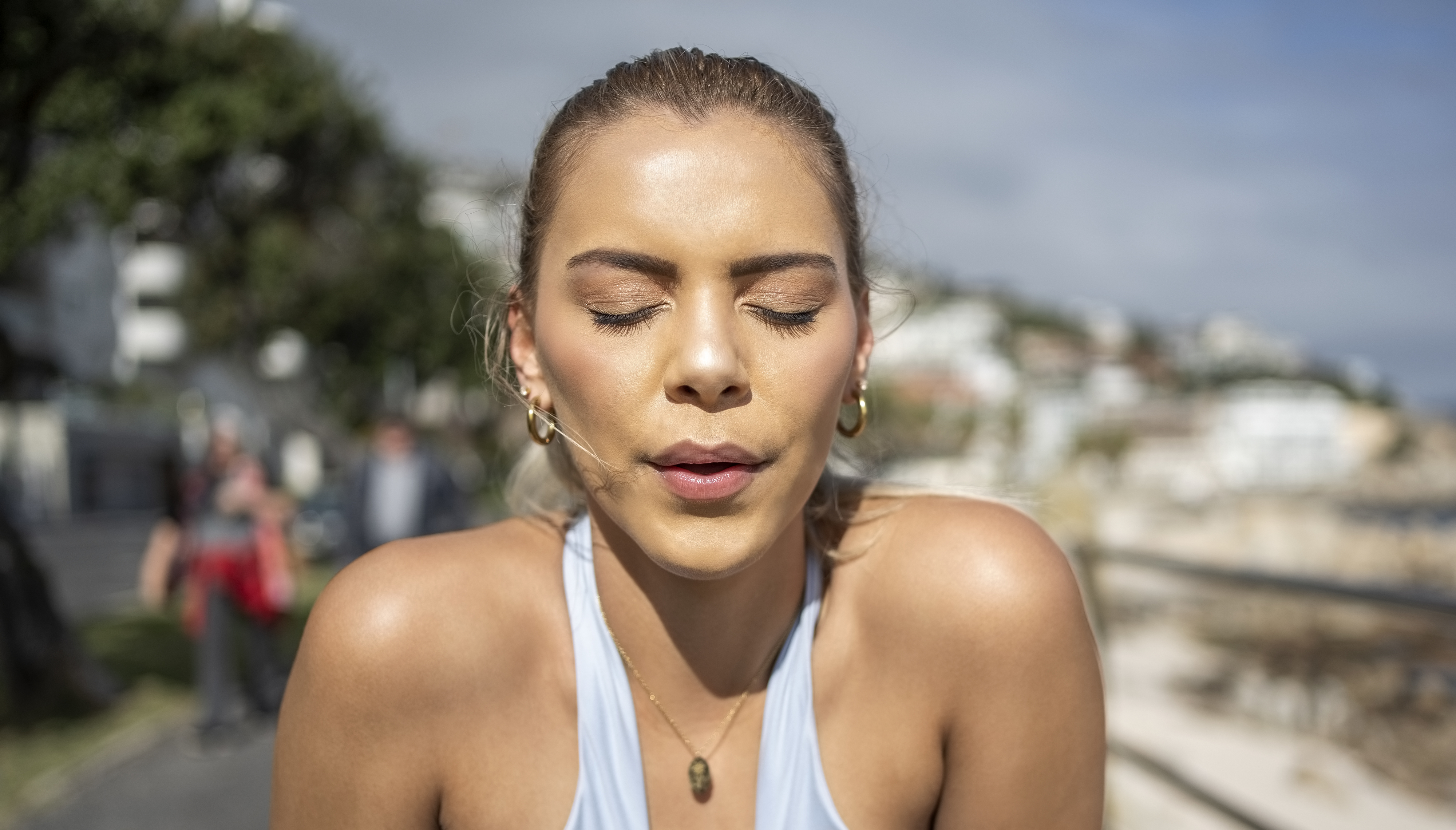 Woman with closed eyes wearing earbuds takes a deep breath outdoors, suggesting relaxation or meditation