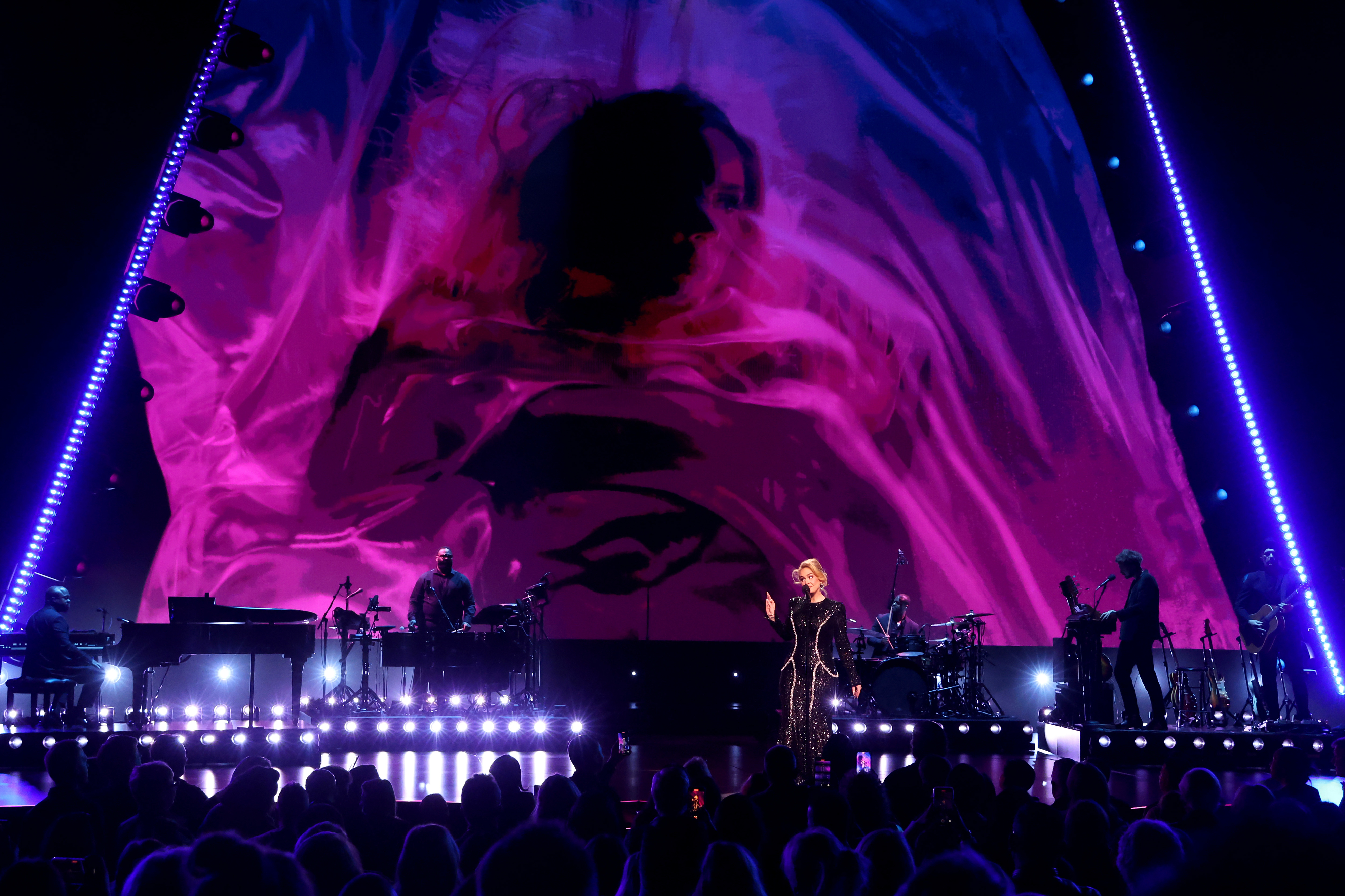 Adele performs on stage with her band, with a large pink abstract backdrop