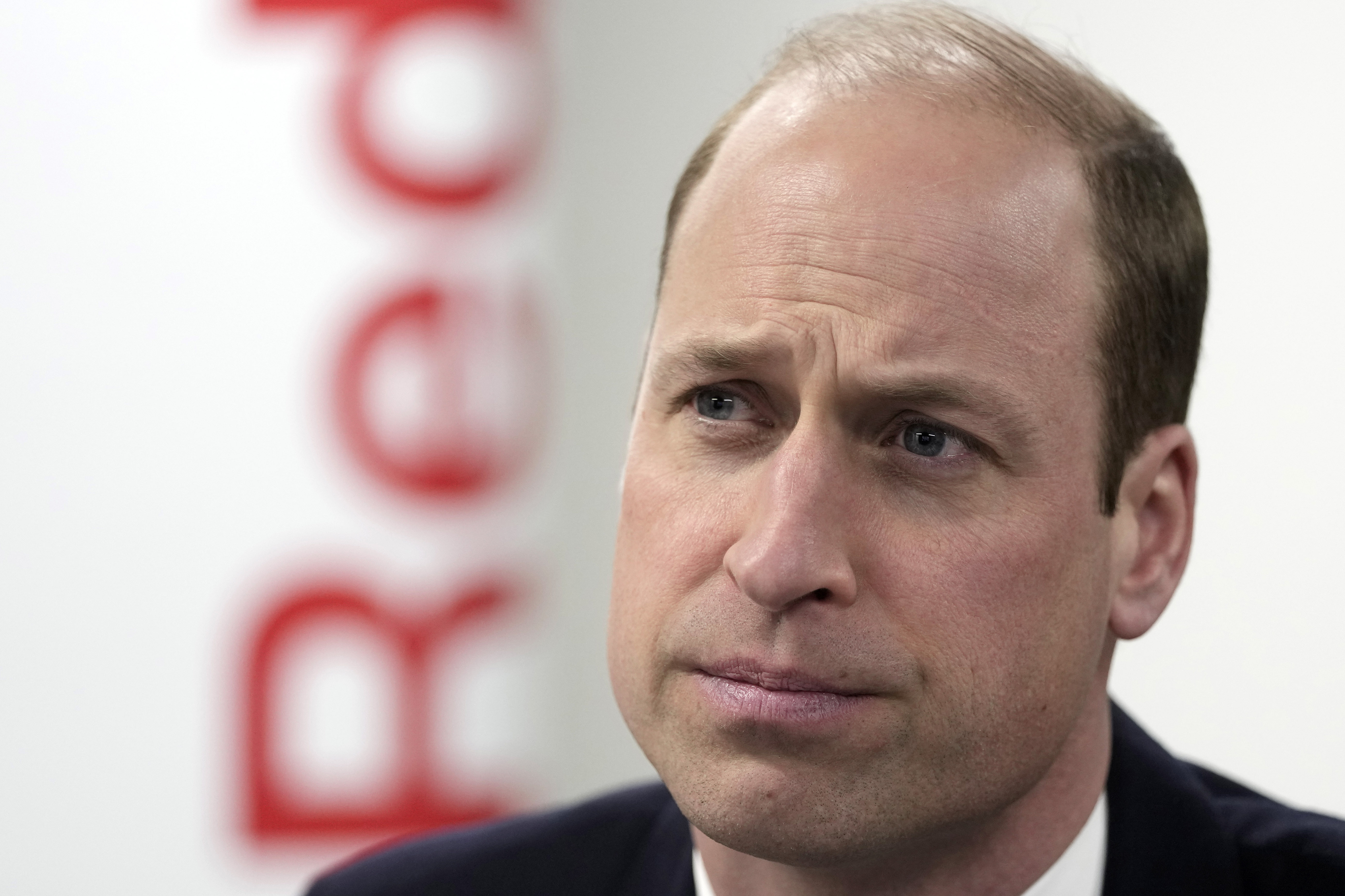 Prince William in a close-up, looking serious, with a blurred logo in the background