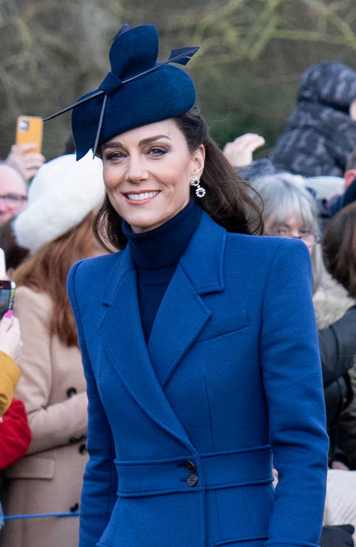 Kate Middleton in a coat and matching hat with feathers smiling as she walks by a crowd