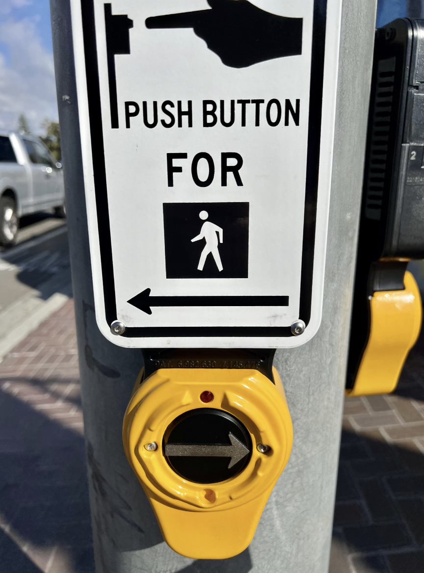 Pedestrian crossing button with instructions "PUSH BUTTON FOR WALK SIGNAL" with the arrow on the sign pointing left in one direction, but the button point right, in the opposite direction