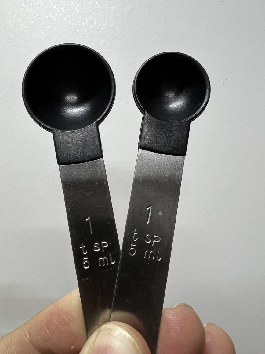 A hand holding two metal 1 teaspoon measuring spoons, with one visibly larger than the other