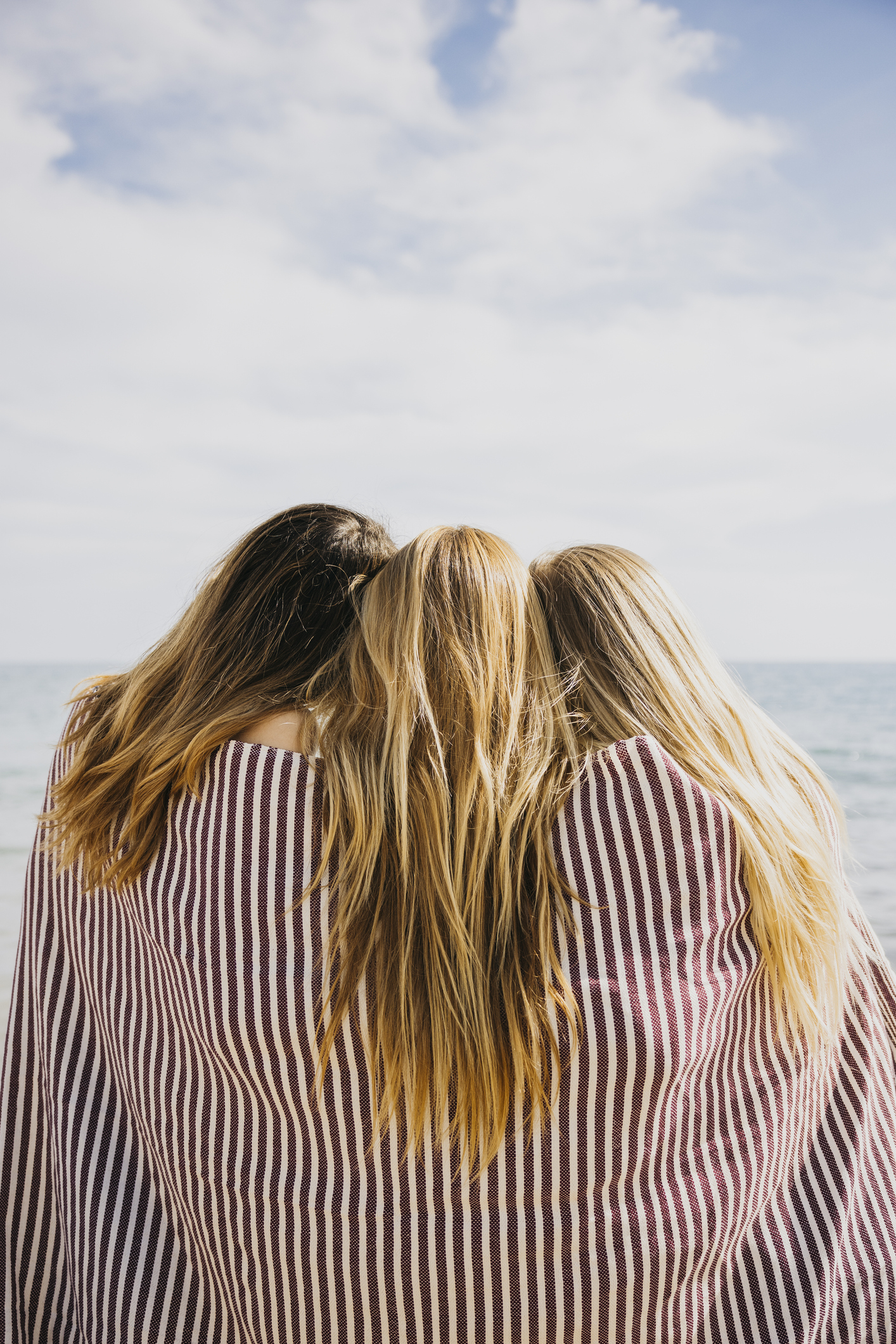 Three friends wrapped inside a blanket as they stand and look out at the beach