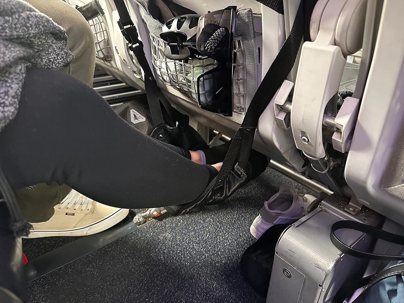 Person wearing sneakers with foot resting on airplane cabin floor near a seated passenger&#x27;s leg, with baggage underneath seat