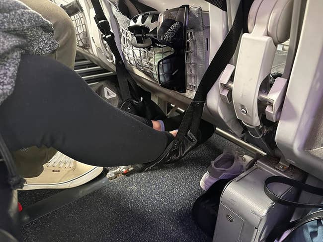 Person wearing sneakers with foot resting on airplane cabin floor near a seated passenger's leg, with baggage underneath seat