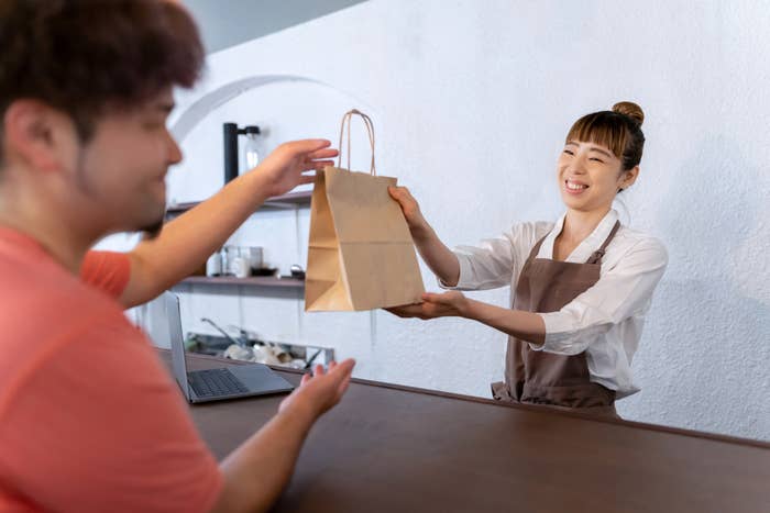 A smiling cashier hands over a paper bag to a customer across the counter