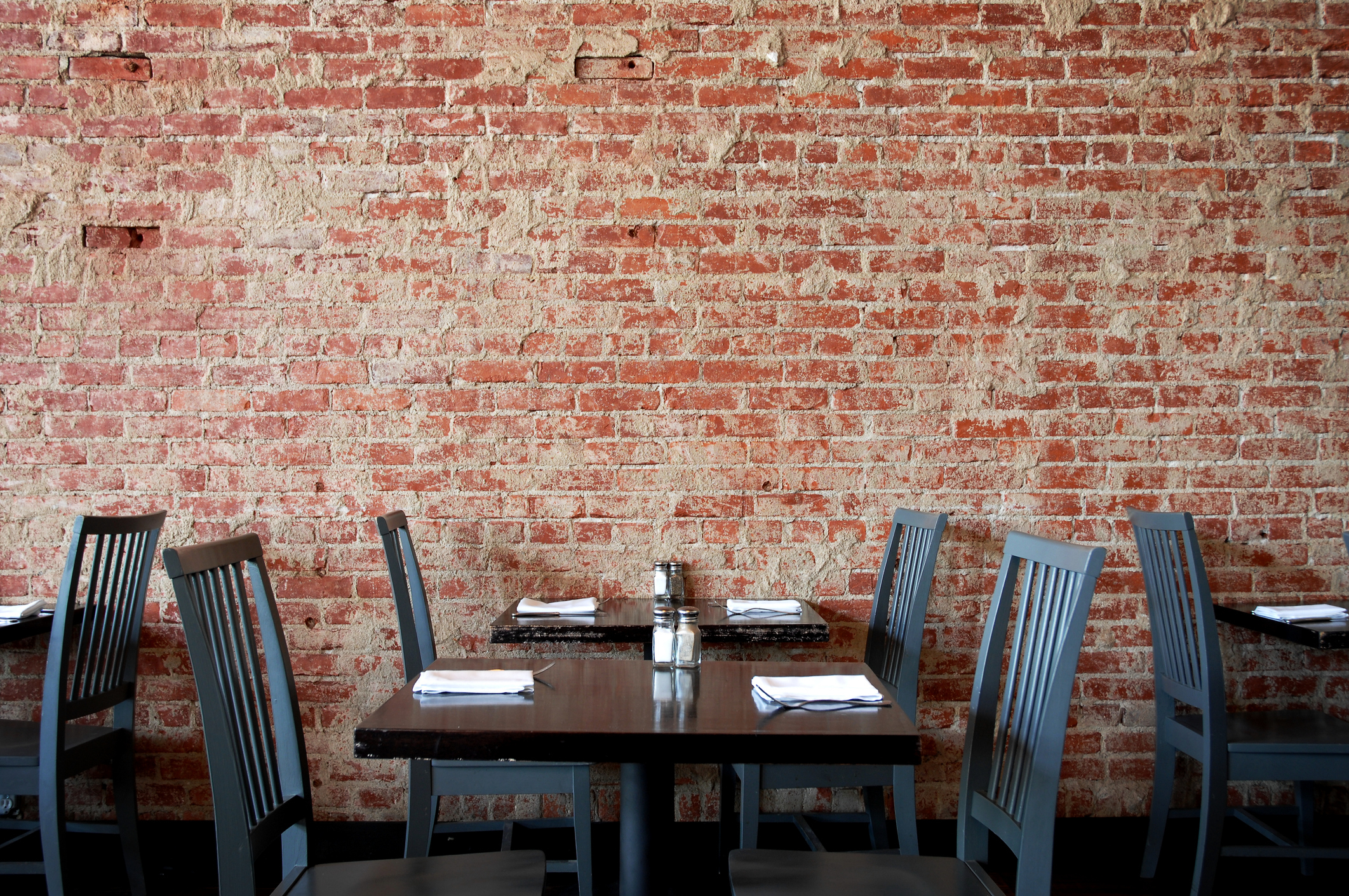 A dining area with empty chairs around tables, set with white plates and napkins, against a brick wall