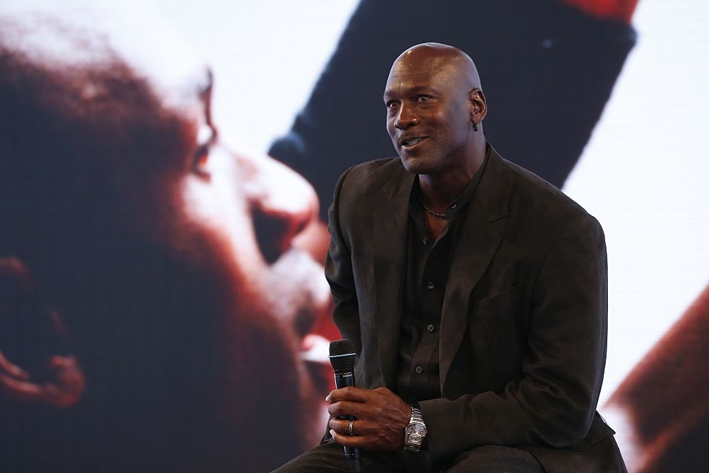 Michael Jordan sits with a microphone in hand, a banner with a basketball player in the background. He wears a suit