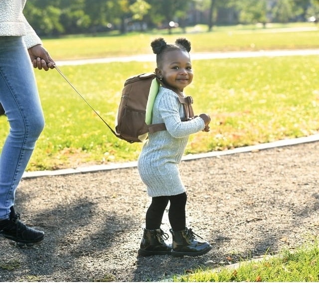 A toddler in a sweater dress and boots with a backpack, held by an adult&#x27;s hand, smiles outdoors