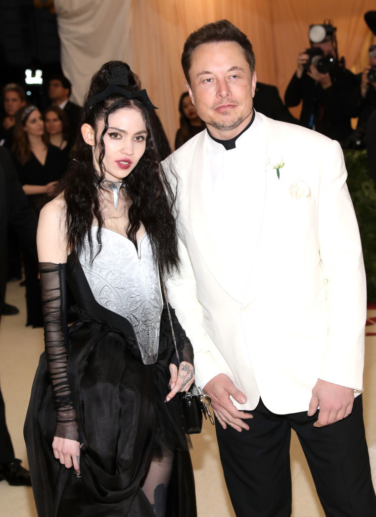 the former couple at an event standing side by side; Elon in a suit with a flower on the lapel, Grimes in a dress with sheer sleeves