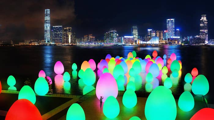 Illuminated egg-shaped sculptures displayed on a waterfront with city skyline in the background at night