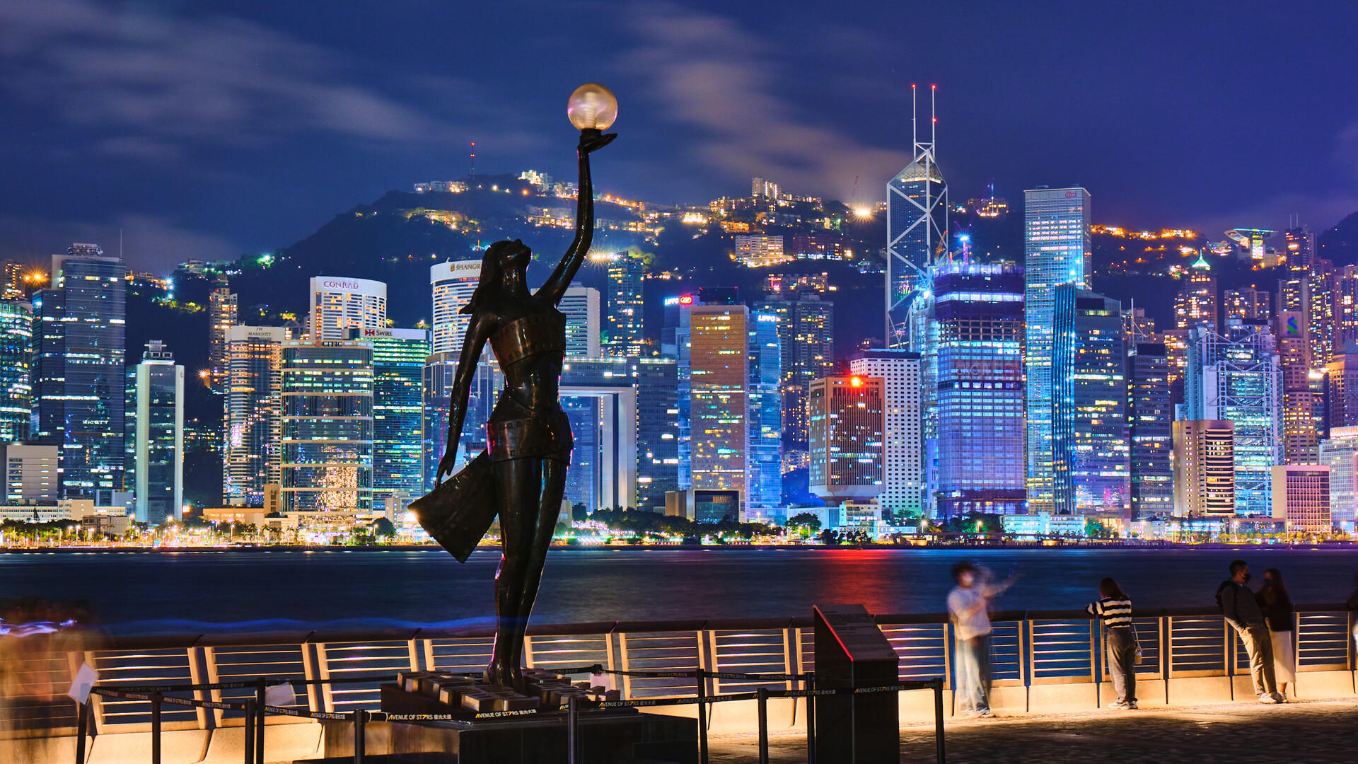 Statue in foreground with city skyline and illuminated buildings at night in the background. People nearby