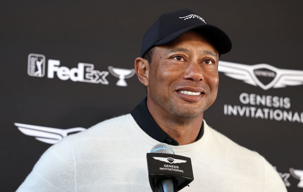 Tiger Woods smiling at Genesis Invitational, wearing a cap and sports jacket, with logos in background