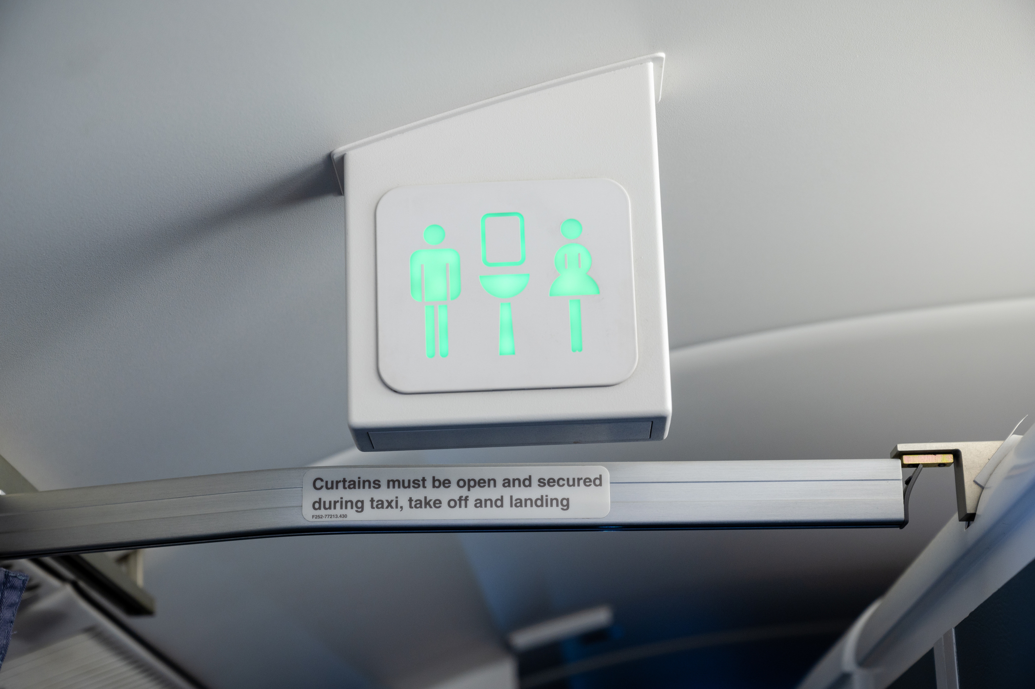 Illuminated airplane lavatory sign with figures, above a text reminder to secure curtains for takeoff/landing