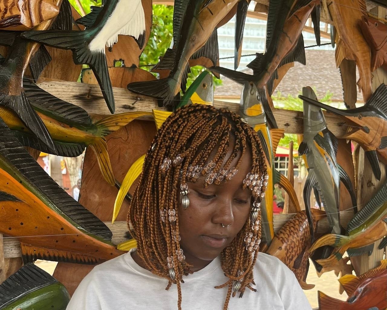 Woman with braided hair and beads, wearing a graphic tee, in front of a background with wooden bird sculptures