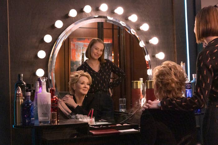 Jean and Hannah in a dressing room with one sitting at a makeup table with a lit mirror, both smiling in a scene from Hacks
