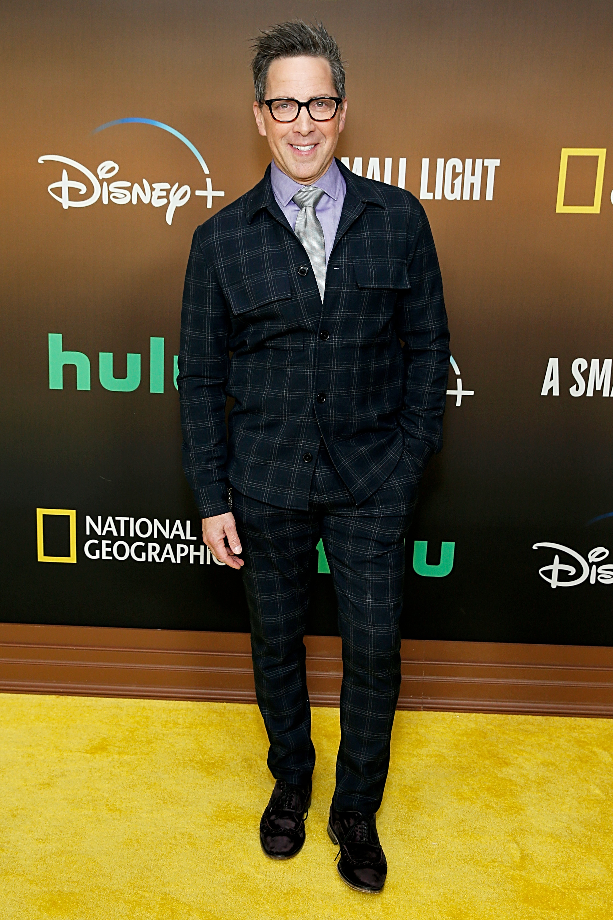 Dan Bucatinsky in plaid suit with glasses on a yellow carpet at a media event