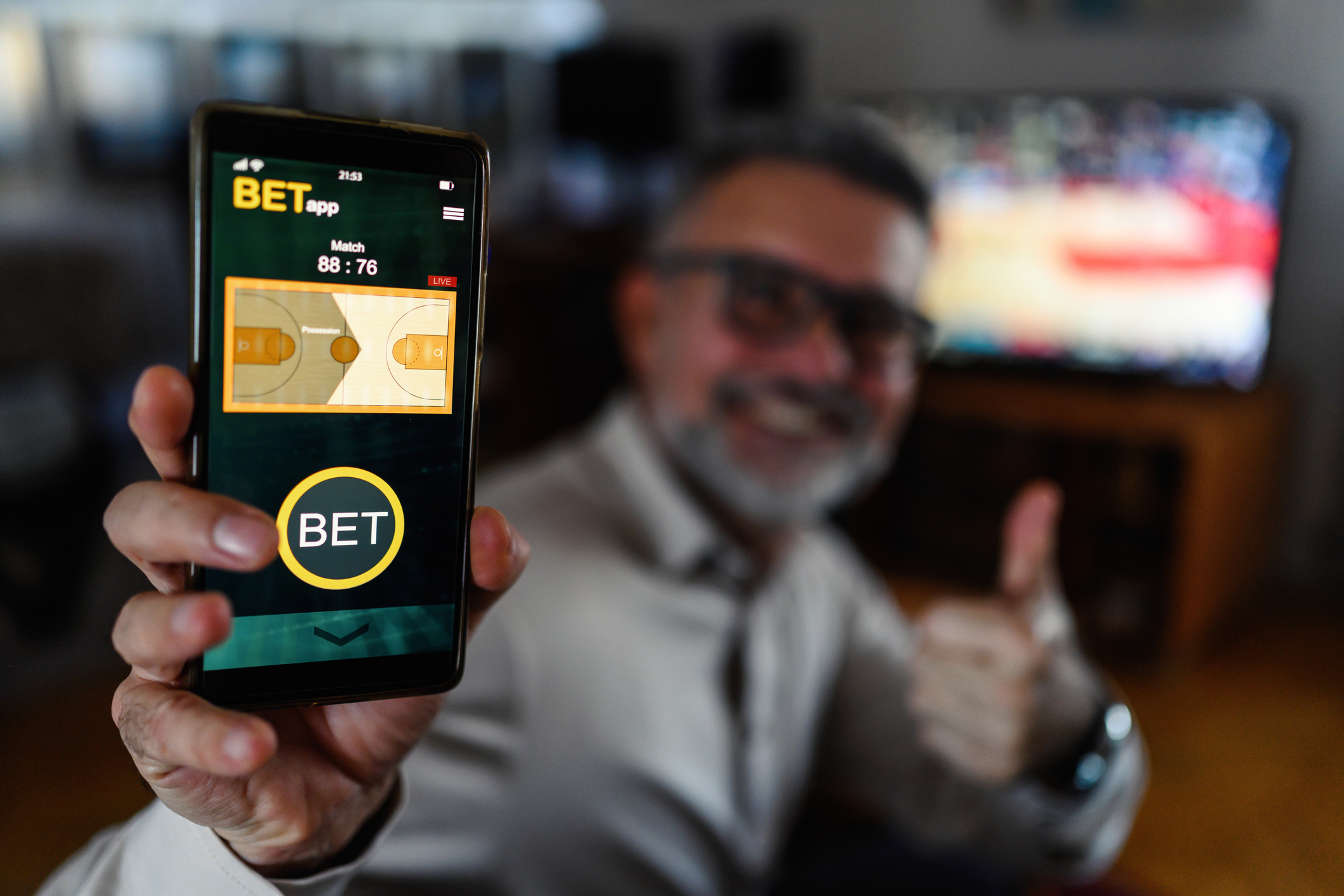 Man showing phone with betting app open, thumb up, blurred TV with sports game in background