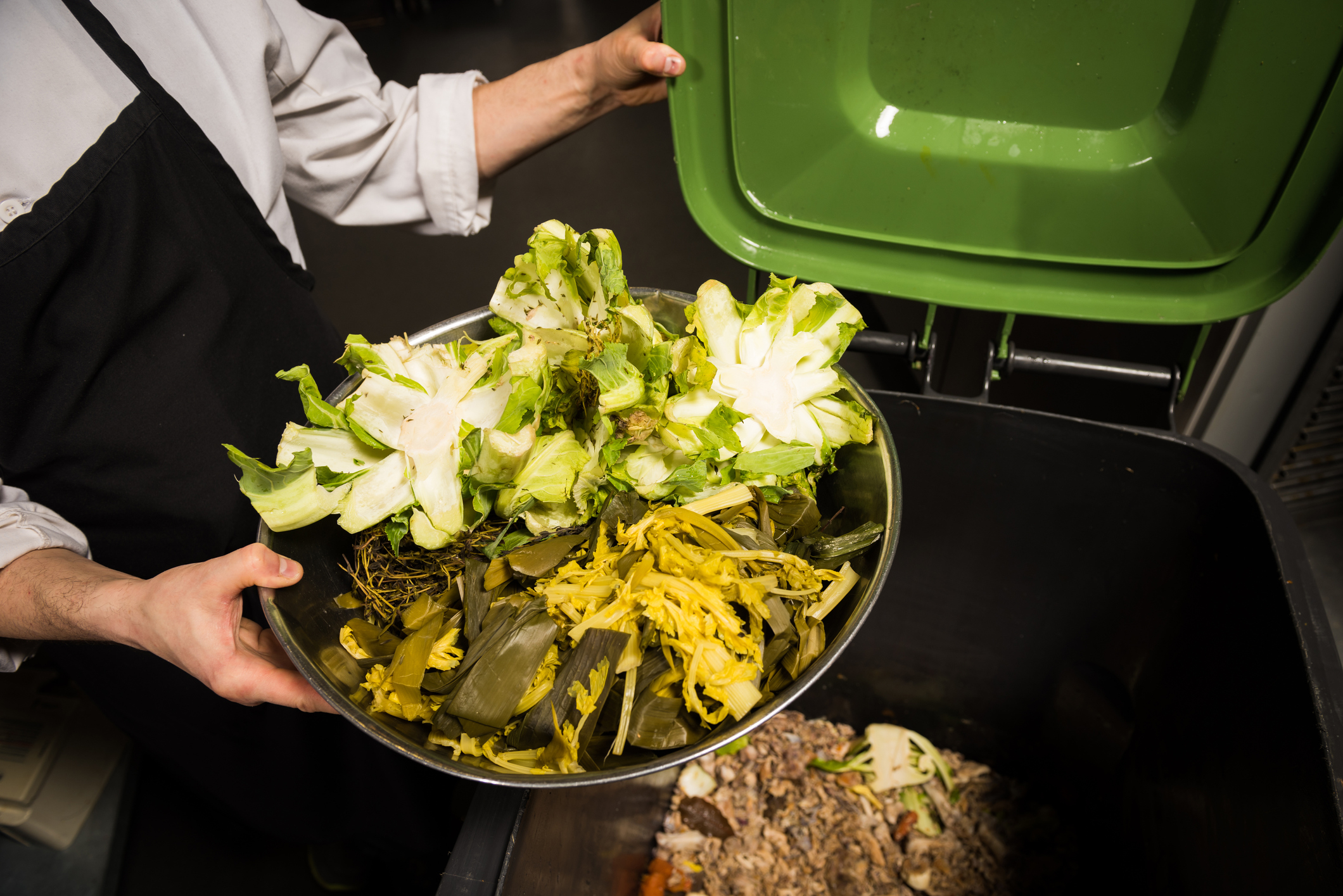 A person is tipping a bowl of food scraps into a green compost bin
