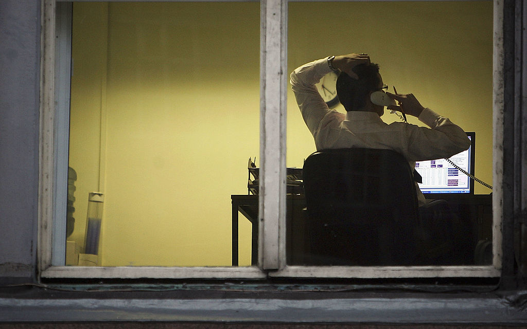 A person is sitting at a desk indoors, using a computer and holding a phone to their ear, visible through a window