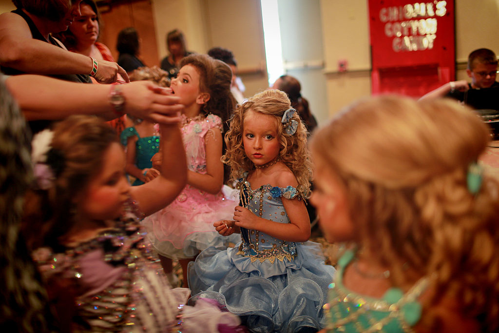 Several children at an event, one in blue dress with curled hair, surrounded by others in glittery costumes