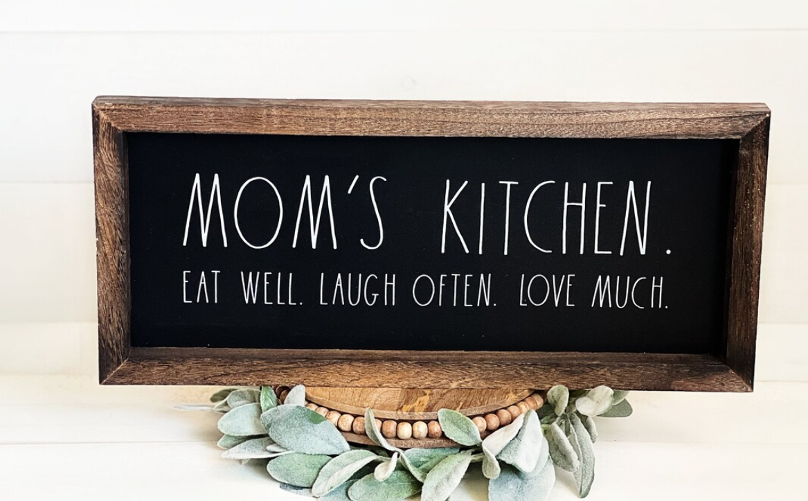 Sign reads &quot;MOM&#x27;S KITCHEN. EAT WELL. LAUGH OFTEN. LOVE MUCH.&quot; on a wooden stand with decorative leaves