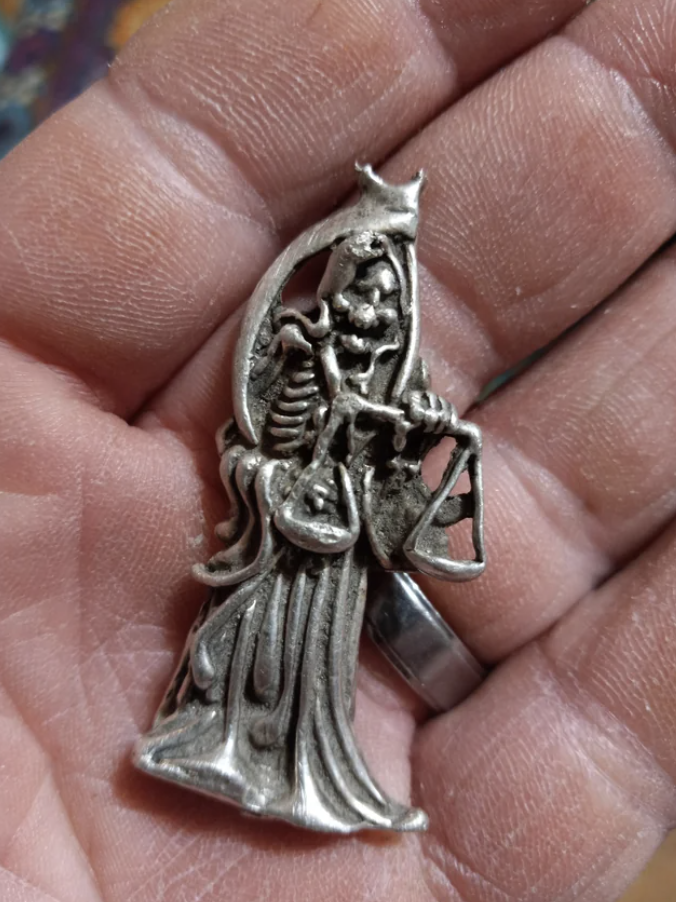 Silver pendant with intricate design of a scary-looking hooded figure holding scales, held in a person&#x27;s hand