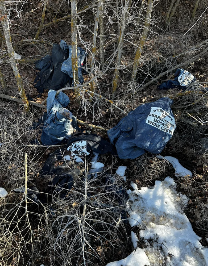 Discarded clothing labeled &quot;Utah Department of Corrections Inmate Work Crew&quot; and trash entangled in barren shrubbery and grass with snow patches on ground
