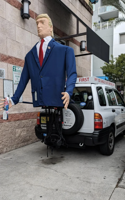 A large Donald Trump figure, with just a head and suit, no pants or bottom torso, on the back of a vehicle