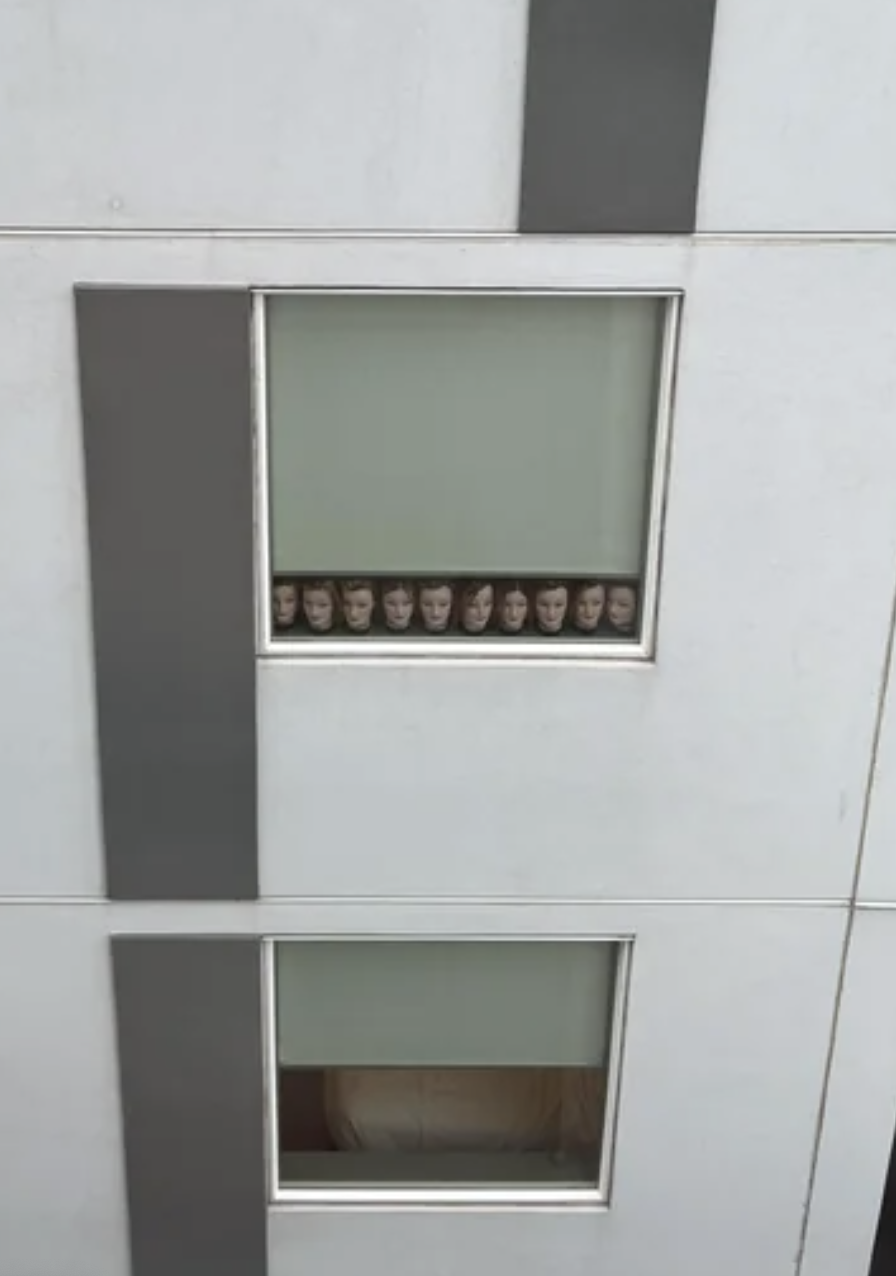 Multiple doll or mannequin heads are seen in a window across from another window, appearing to be in a line