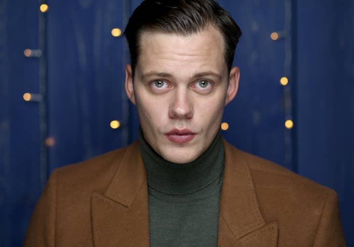 Bill Skarsgård in a brown coat over a green turtleneck, with a focused expression, standing in front of a blue background with soft lights