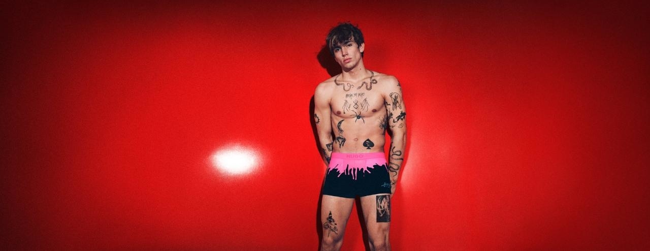Shirtless man with tattoos wearing patterned shorts against red background