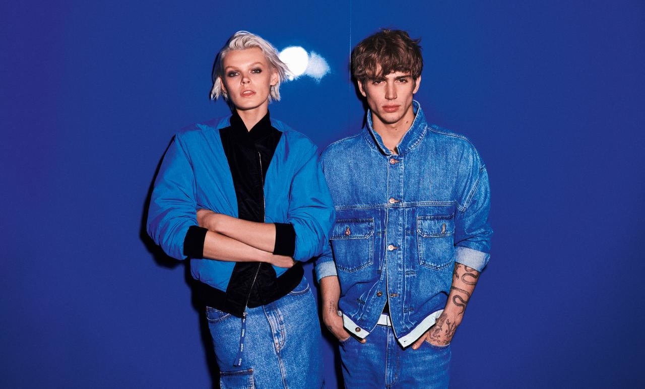 Two models posing, one in a blue jacket and black top, the other in a denim outfit, with hands in pockets