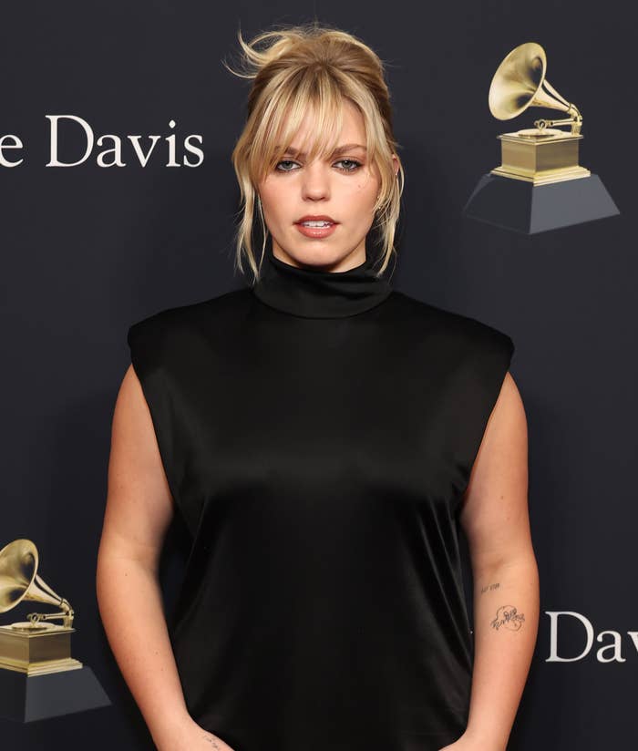 Reneé Rapp in a sleeveless black dress stands against backdrop with text; has a tattoo on left arm