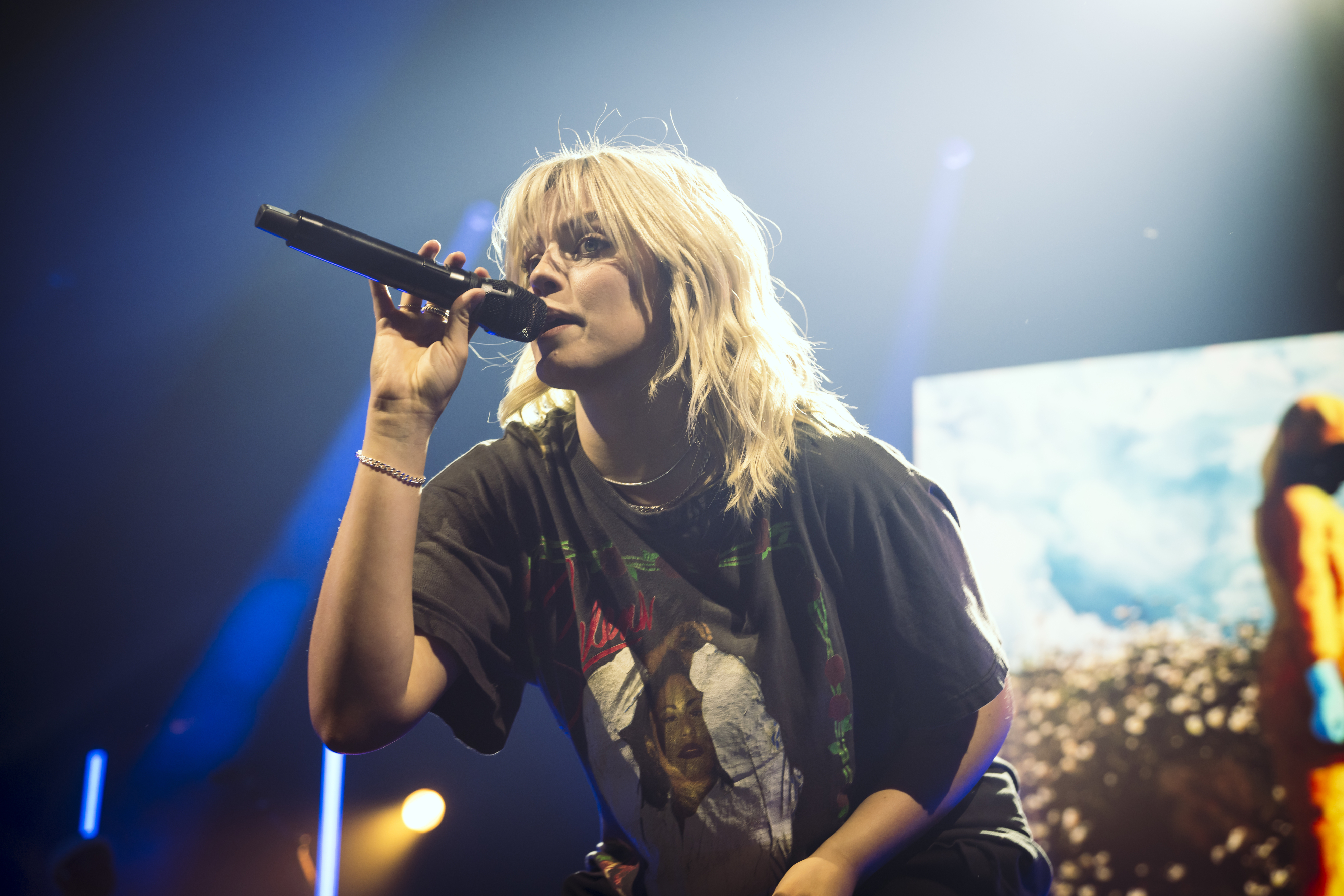 Reneé Rapp with microphone onstage wearing a graphic T-shirt
