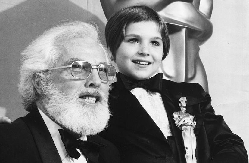 Elderly man and young boy in tuxedos, holding an Oscar statue, smiling
