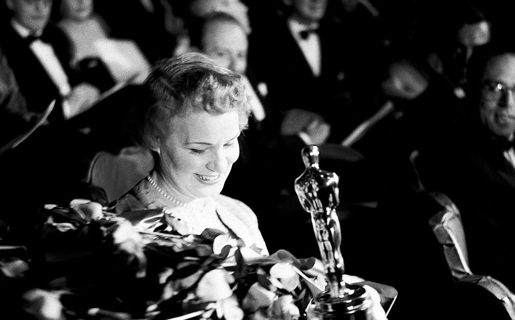 Woman holding an Oscar amidst a crowd, surrounded by flowers, expressing joy