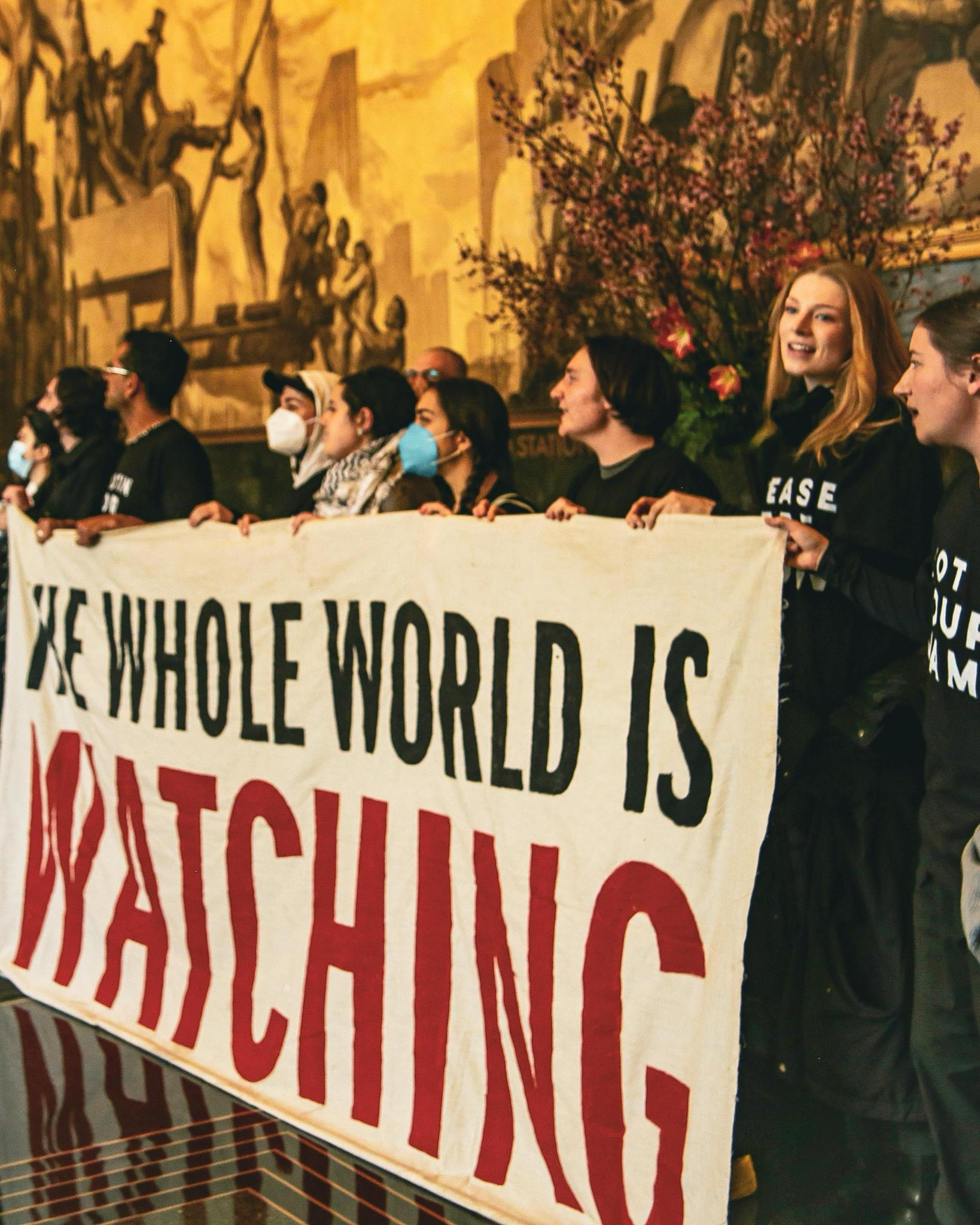 Group of individuals holding a banner reading &quot;THE WHOLE WORLD IS WATCHING&quot; at an event