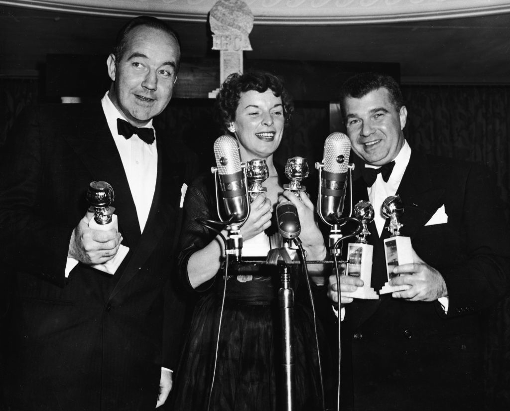 Three people are smiling, holding microphones and awards at an event