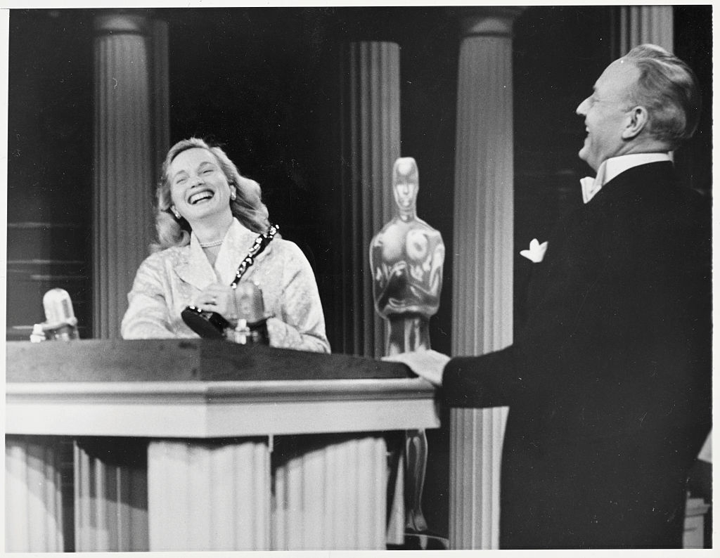 Woman laughing beside podium with Oscar statuette; man smiling at her. Both are dressed in formal attire