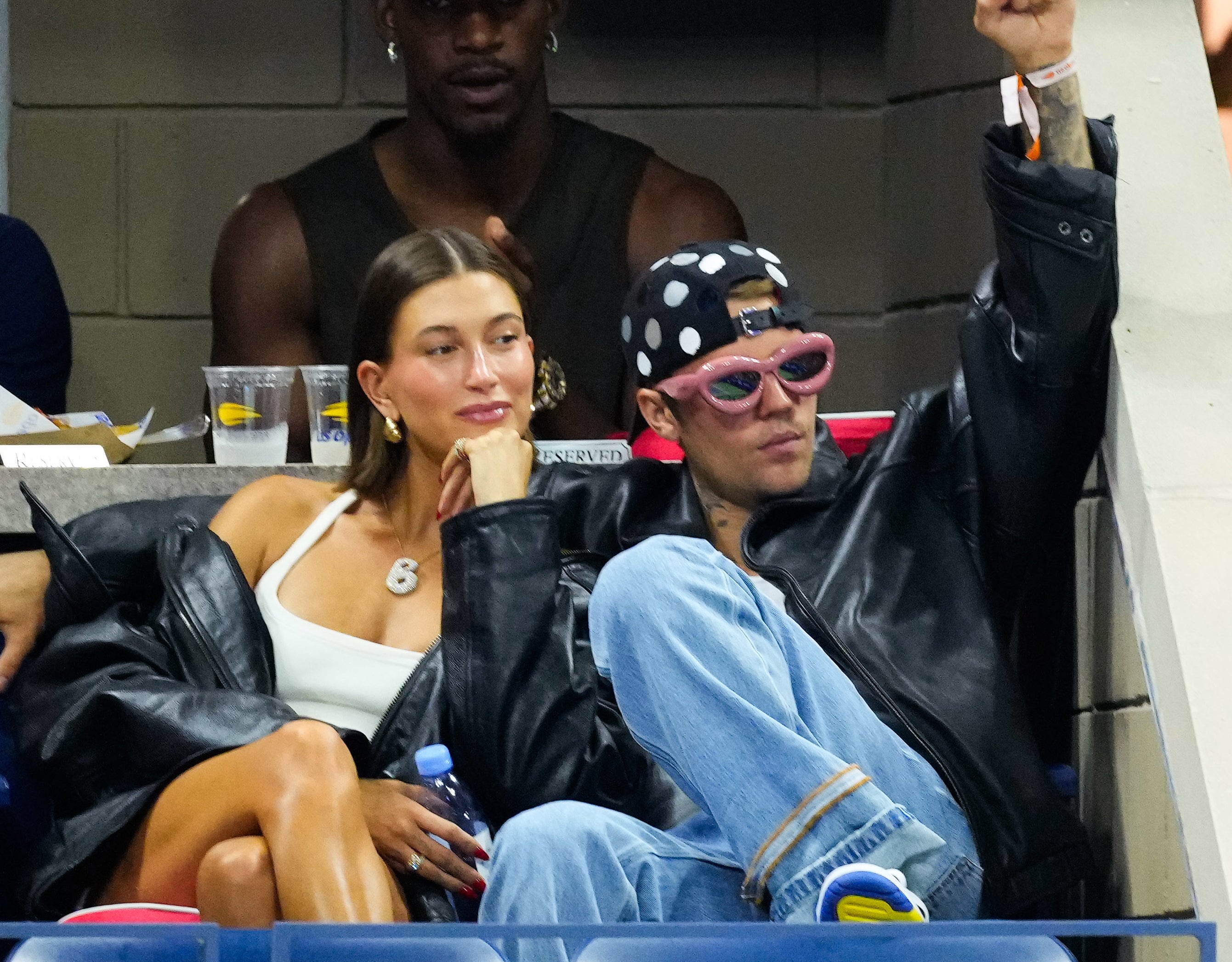 Justin Bieber wearing a leather jacket and cap seated beside Hailey Bieber at a sports event