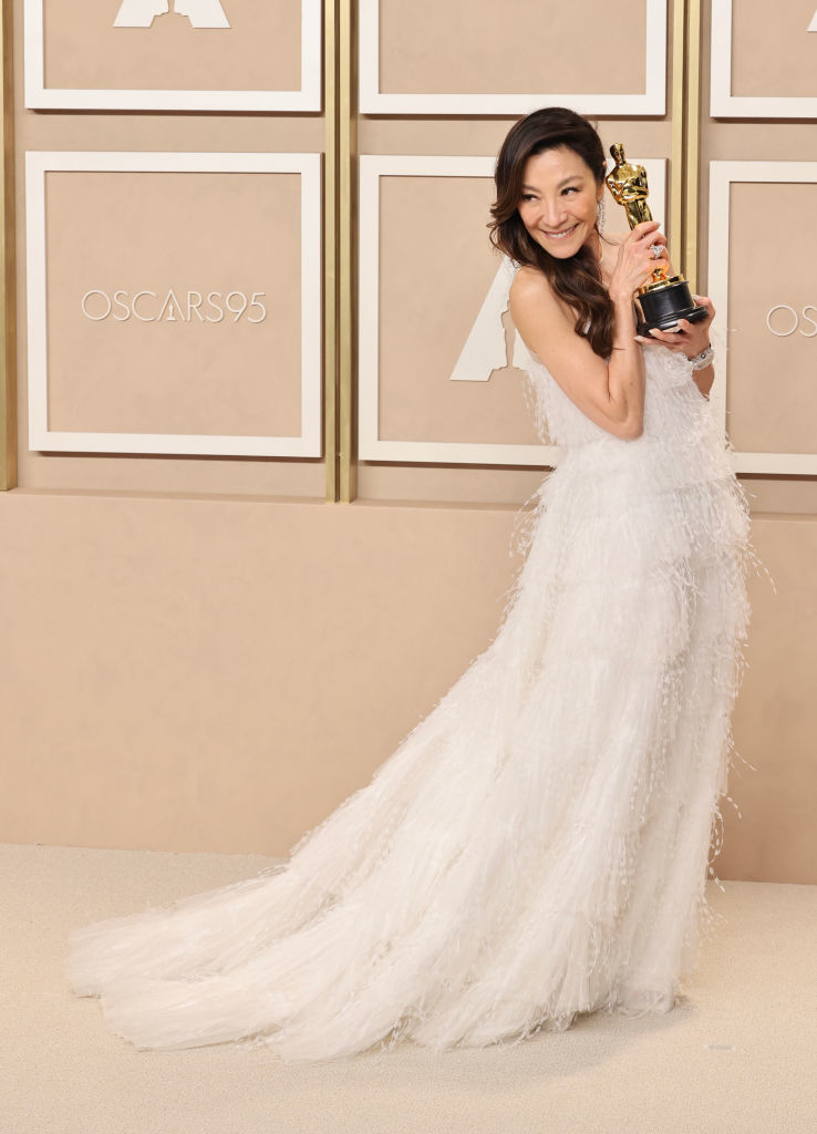 Michelle holding an Oscar statuette, wearing an elegant gown with a long, feathered skirt at an award event