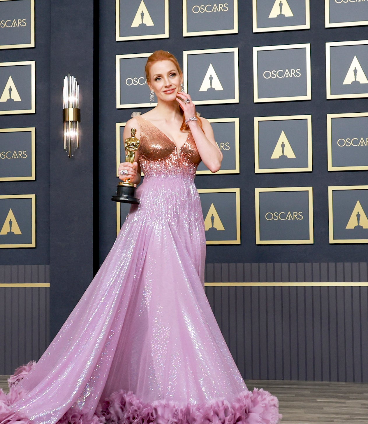 Jessica in a sparkling gown with a deep neckline and voluminous skirt, holding an award statue, poses at the Oscars event