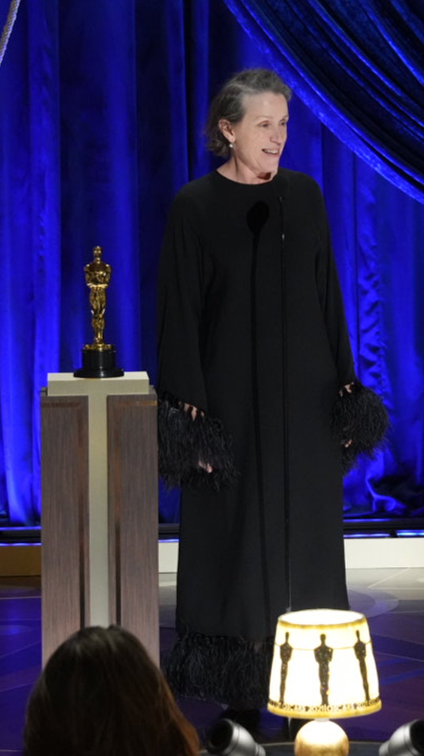 Frances in a dark outfit with fringe details stands at a podium with a trophy, addressing an audience