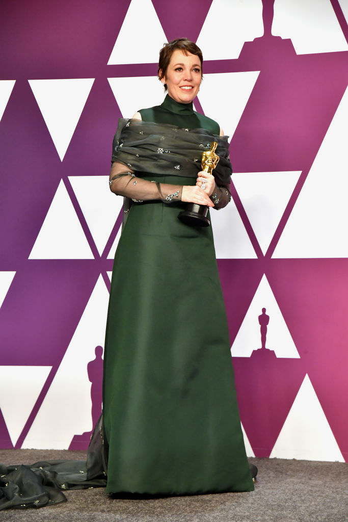 Olivia Colman in a long-sleeve gown, holding an Oscar trophy at an awards backdrop
