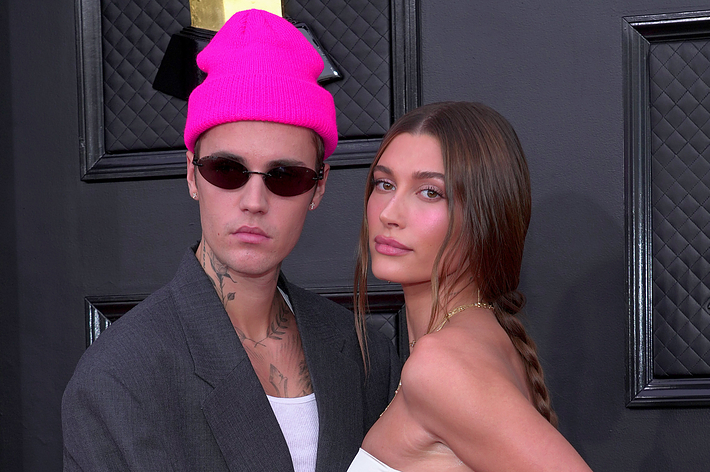 Justin Bieber in oversized gray suit and pink beanie, Hailey Bieber in white dress with side slit, both posing at an event