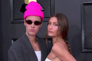 Justin Bieber in oversized gray suit and pink beanie, Hailey Bieber in white dress with side slit, both posing at an event