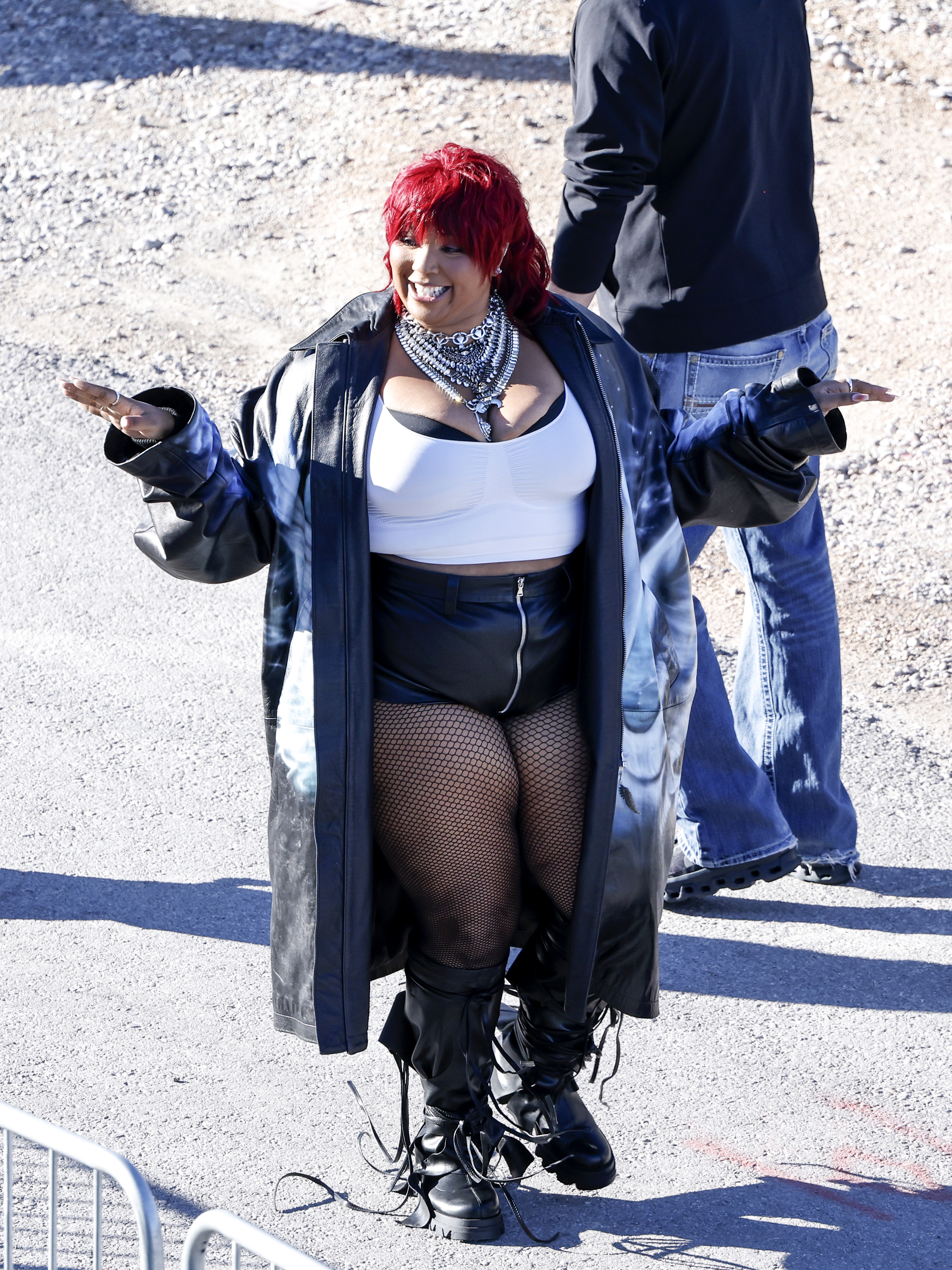 Lizzo is wearing a top, jacket, fishnet tights, and boots as she smiles and gestures while walking outside at an event