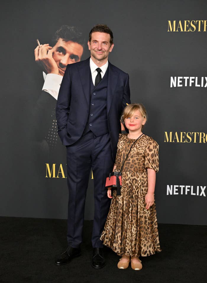 Bradley Cooper in a suit standing with his young daughter, Lea, in a leopard-print dress at a Netflix event