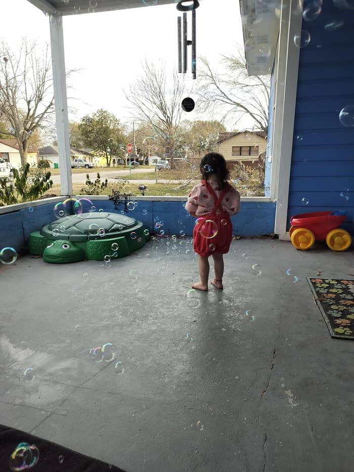Child in a wig and backpack plays with bubbles on a porch, near toy cars